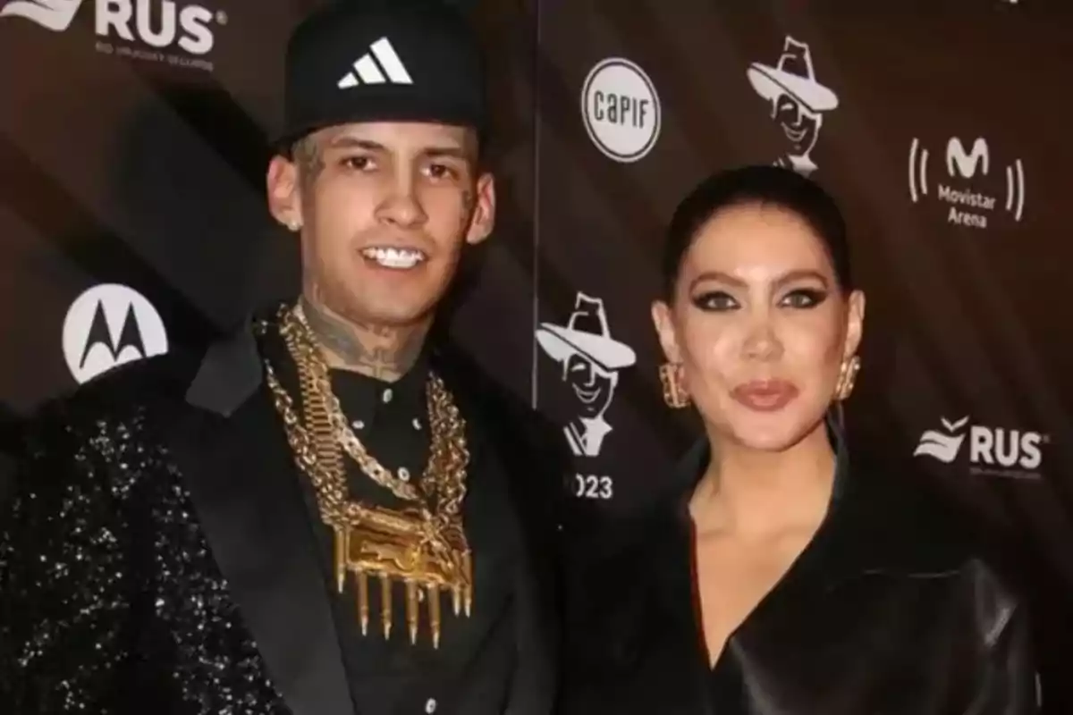 Two people pose together at an event with a backdrop of logos, both wearing elegant clothing and striking accessories.