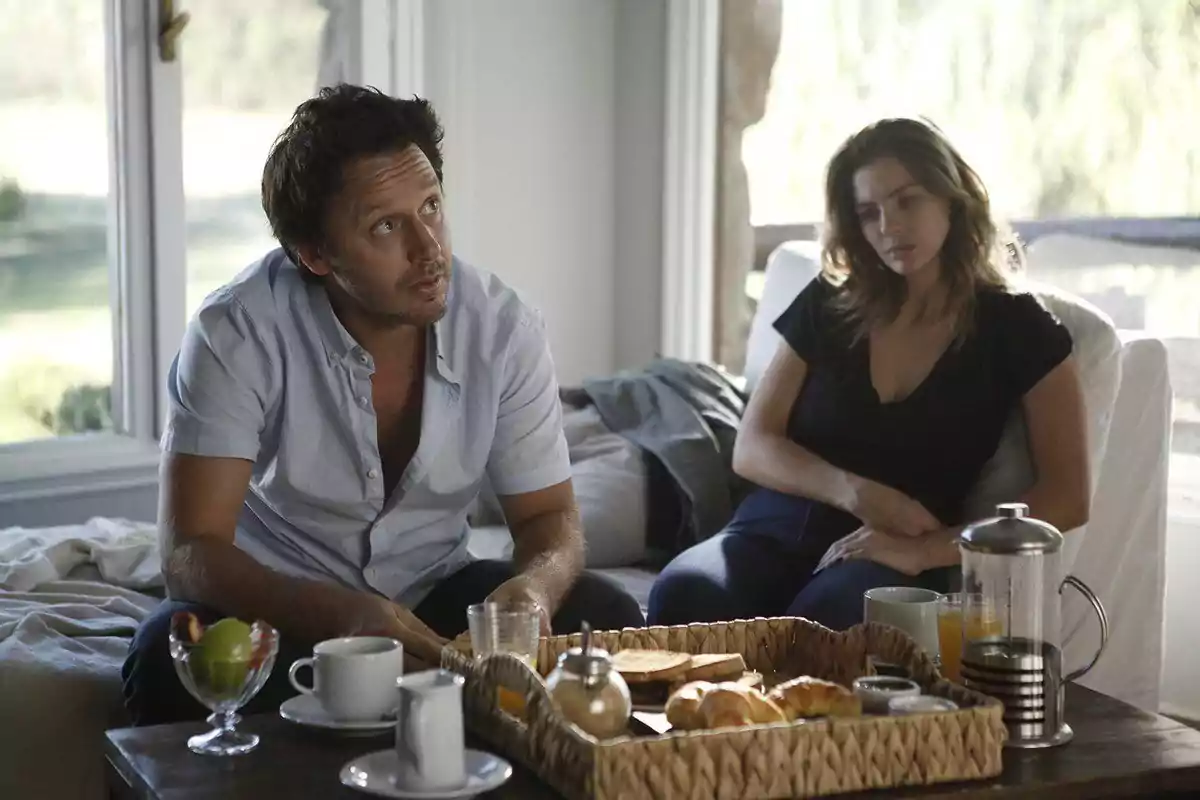 Una pareja sentada en una sala con expresión seria mientras tienen un desayuno servido en una mesa frente a ellos Una pareja sentada en una sala con expresión seria mientras tienen un desayuno servido en una mesa frente a ellos
