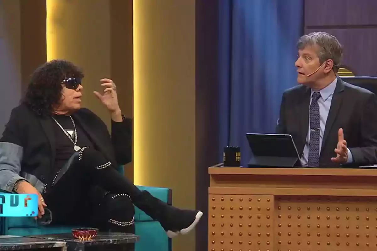 Two people are talking on a television set; one of them is wearing dark glasses and the other is sitting behind a desk with a tablet. Two people are talking on a television set; one of them is wearing dark glasses and the other is sitting behind a desk with a tablet.
