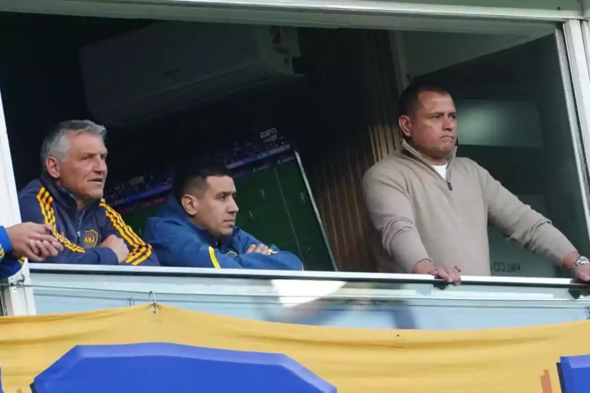 Three men are watching a soccer match from a box in a stadium; two of them are wearing blue and yellow sportswear, and the other is wearing a beige sweater.