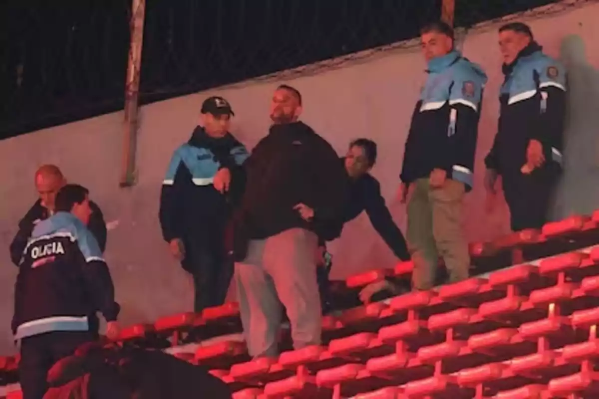 Varios policías y personas están en las gradas de un estadio, algunos de pie y otros agachados, en un ambiente nocturno. Varios policías y personas están en las gradas de un estadio, algunos de pie y otros agachados, en un ambiente nocturno.