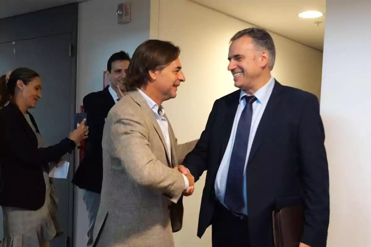 Dos hombres vestidos de traje se saludan con un apretón de manos y sonríen mientras otras personas observan en el fondo