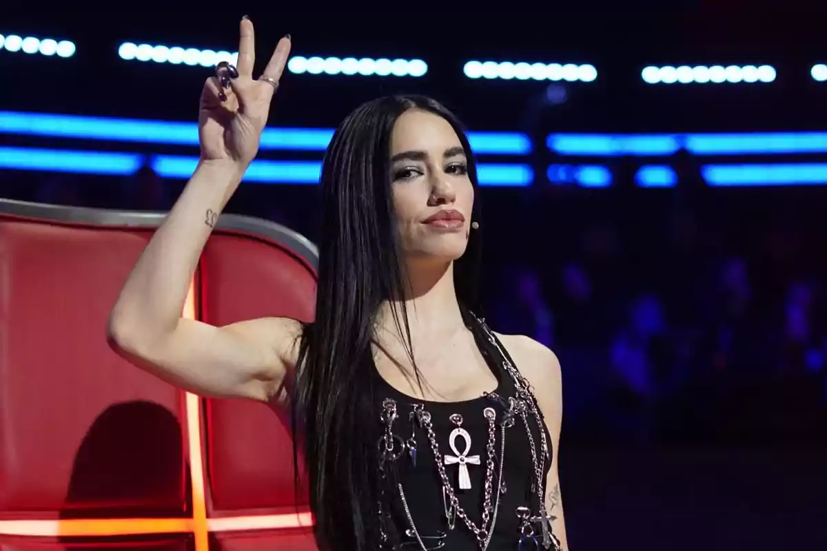 Mujer de cabello largo y oscuro haciendo el signo de la paz con la mano mientras está sentada en una silla roja en un set iluminado