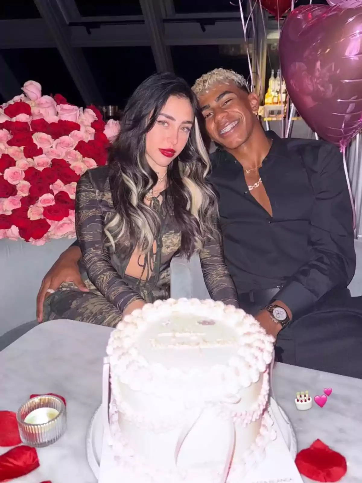 Couple sitting embraced, celebrating with a decorated white cake, surrounded by heart-shaped balloons and a large bouquet of red and pink roses Couple sitting embraced, celebrating with a decorated white cake, surrounded by heart-shaped balloons and a large bouquet of red and pink roses