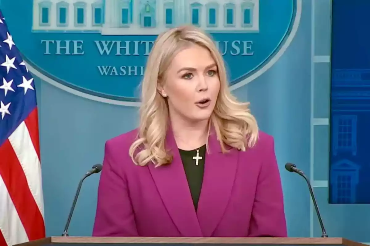 Blonde woman in a fuchsia blazer speaking at a podium in front of a White House backdrop and a United States flag