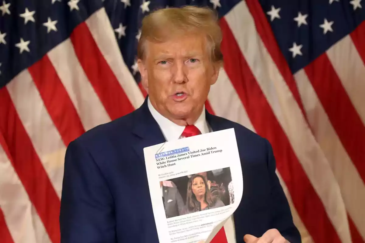 Blond-haired man in a blue suit and red tie holds a printed sheet in front of several United States flags