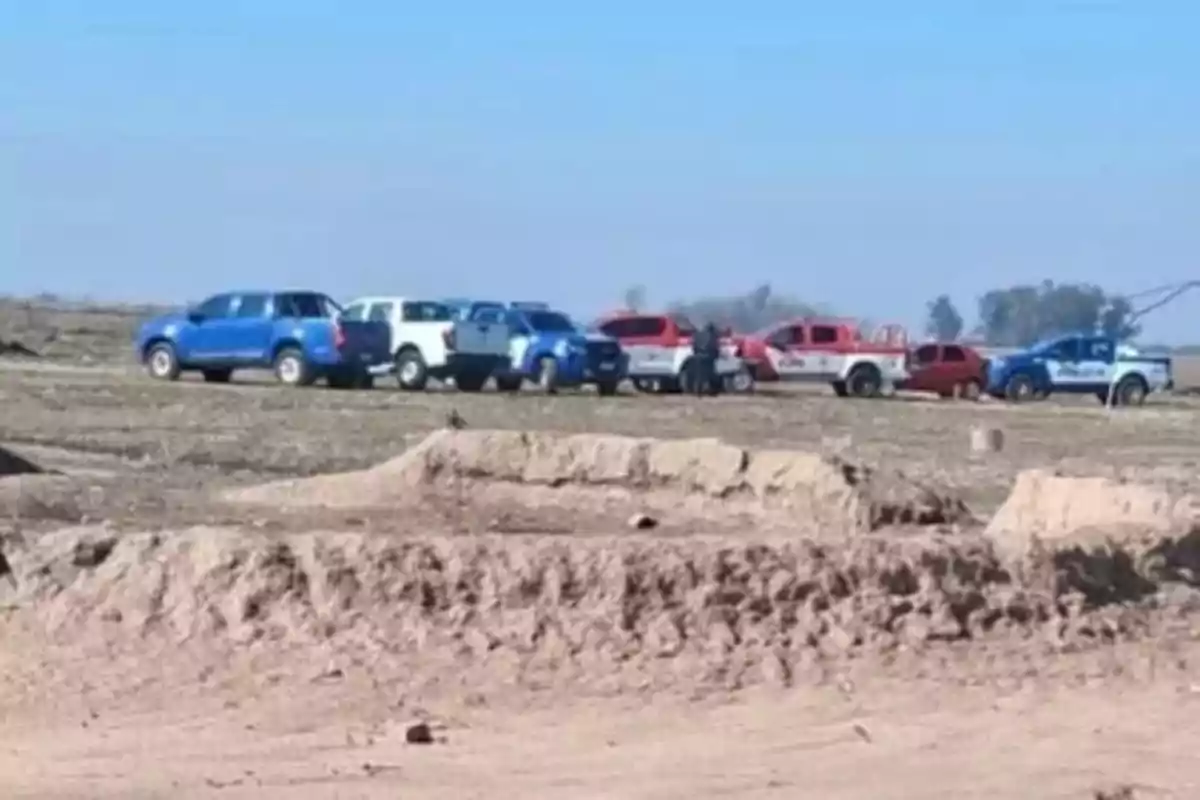 Varias camionetas de diferentes colores y personas se encuentran reunidas en un terreno árido bajo un cielo despejado. Varias camionetas de diferentes colores y personas se encuentran reunidas en un terreno árido bajo un cielo despejado.