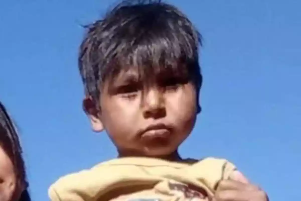 Niño con cabello oscuro y expresión seria mirando a la cámara con el cielo azul de fondo Niño con cabello oscuro y expresión seria mirando a la cámara con el cielo azul de fondo