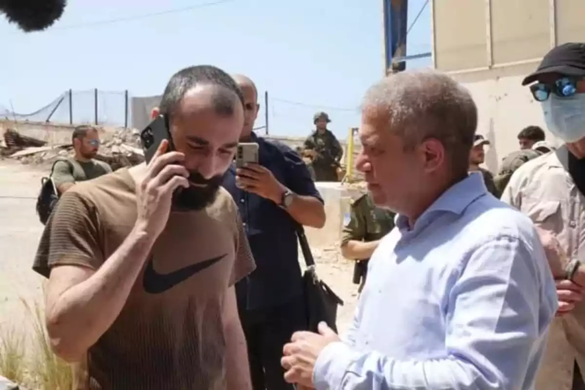 Two men are talking outdoors while one of them is on the phone, and several people, some in military uniforms, are in the background.