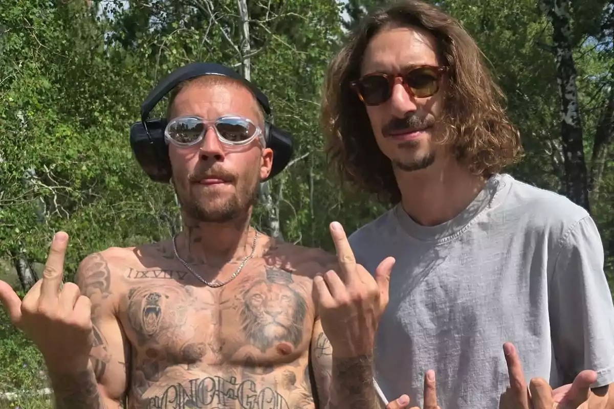 Two men pose outdoors showing their middle fingers to the camera; one of them is jerseyless and wearing glasses and headphones, while the other is wearing a gray t-jersey and sunglasses.