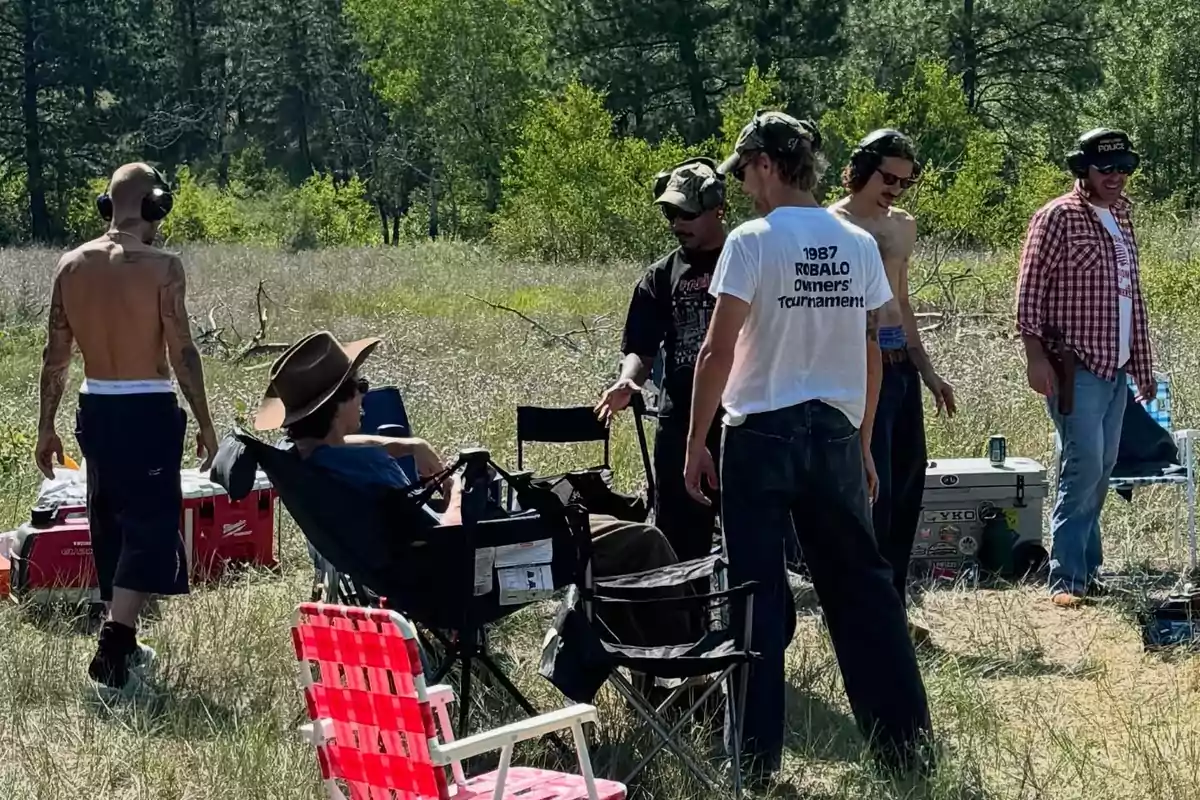 A group of people wearing hearing protection are outdoors in a field surrounded by trees; some are standing and others are sitting on folding chairs, enjoying a sunny day. A group of people wearing hearing protection are outdoors in a field surrounded by trees; some are standing and others are sitting on folding chairs, enjoying a sunny day.
