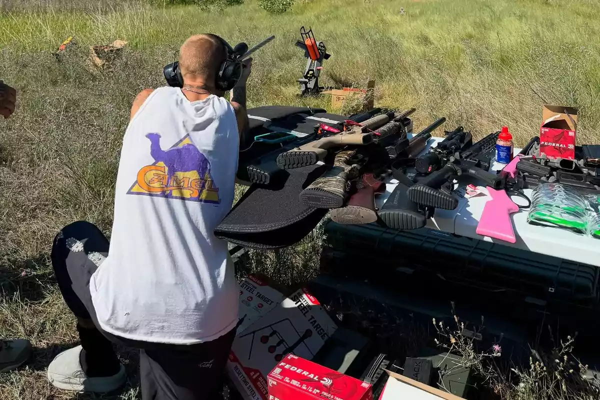 Person wearing protective earmuffs shooting a rifle in an open field next to a table full of guns and shooting equipment Person wearing protective earmuffs shooting a rifle in an open field next to a table full of guns and shooting equipment