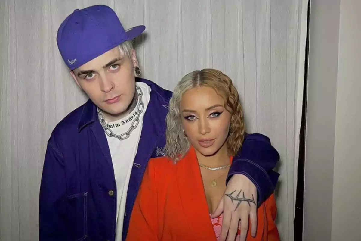 Two people pose together in front of a white curtain; one is wearing a blue cap and a silver chain, the other is wearing an orange jacket and has wavy blonde hair.