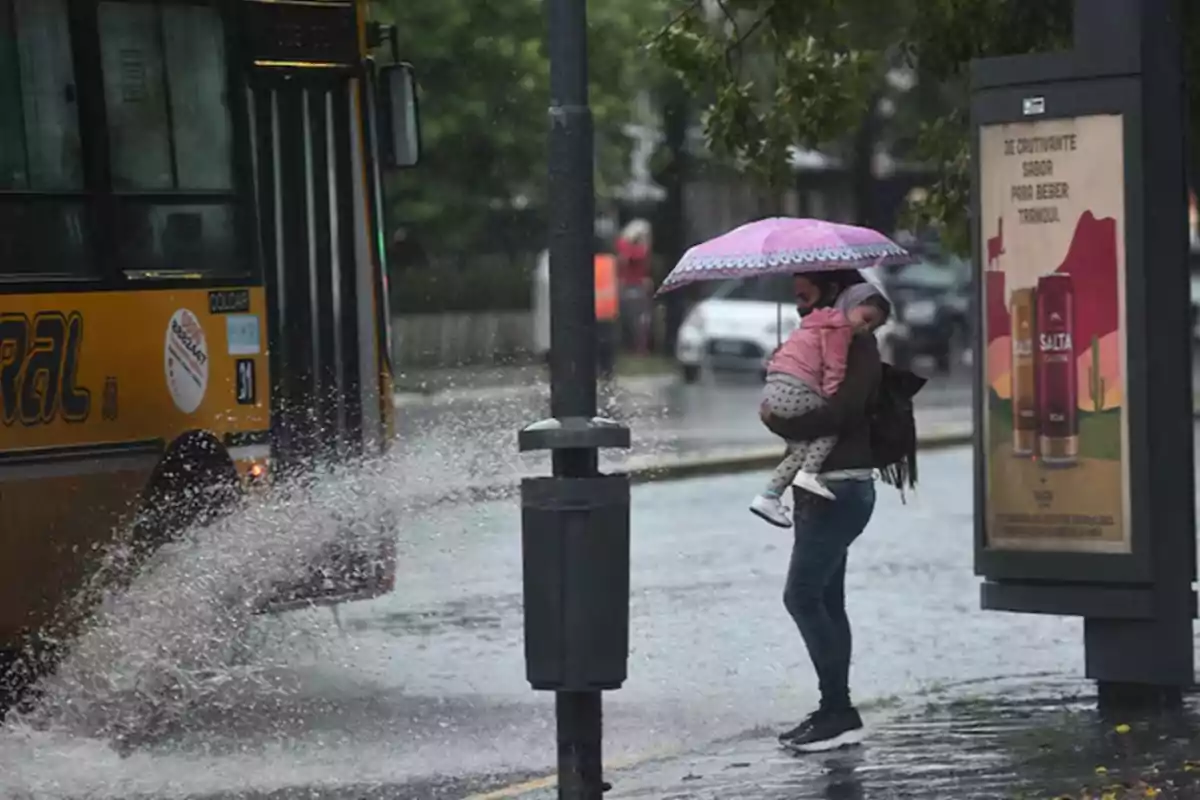 A woman holds a girl under a pink umbrella as a bus passes by and splashes water on a rainy street. A woman holds a girl under a pink umbrella as a bus passes by and splashes water on a rainy street.