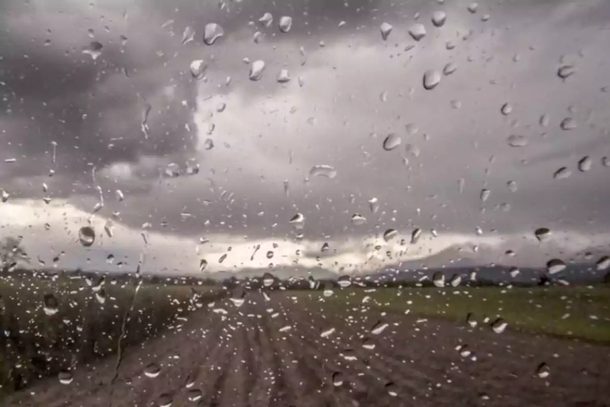 Gotas de lluvia en una ventana con un paisaje nublado y un camino de tierra al fondo Gotas de lluvia en una ventana con un paisaje nublado y un camino de tierra al fondo