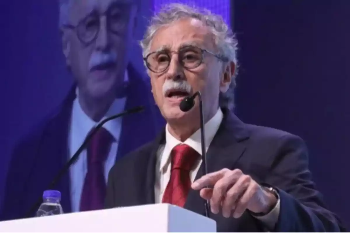 Hombre de cabello canoso y bigote usando gafas y traje oscuro con corbata roja hablando en un podio con micrófono y fondo azul con su imagen proyectada detrás Hombre de cabello canoso y bigote usando gafas y traje oscuro con corbata roja hablando en un podio con micrófono y fondo azul con su imagen proyectada detrás