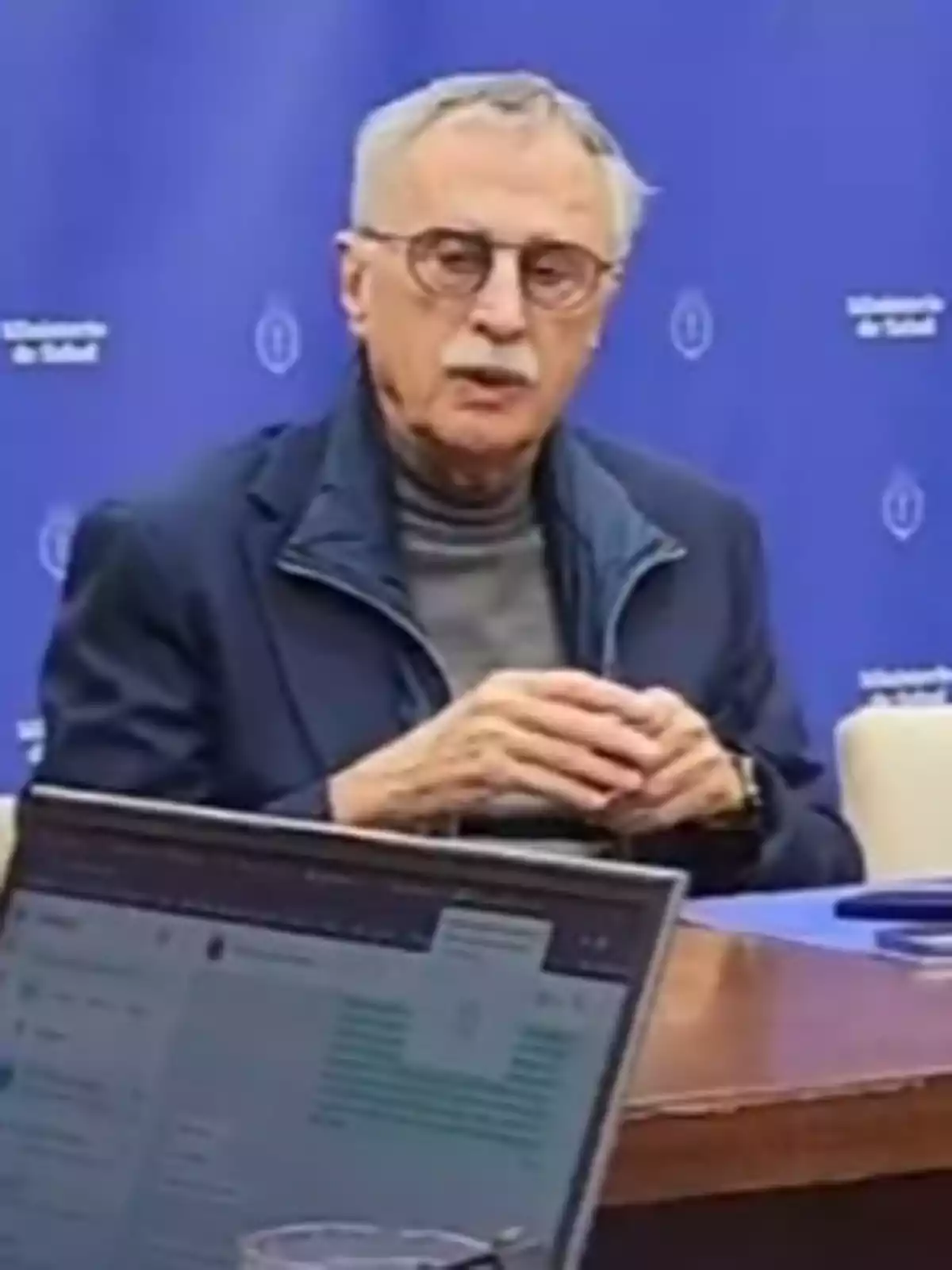 Hombre mayor con gafas y bigote sentado frente a una mesa con una laptop, fondo azul con logotipos institucionales Hombre mayor con gafas y bigote sentado frente a una mesa con una laptop, fondo azul con logotipos institucionales