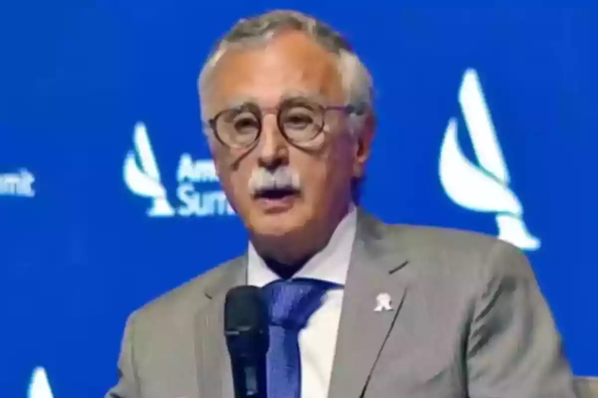 Hombre de cabello canoso y bigote con gafas hablando en un evento frente a un fondo azul Hombre de cabello canoso y bigote con gafas hablando en un evento frente a un fondo azul