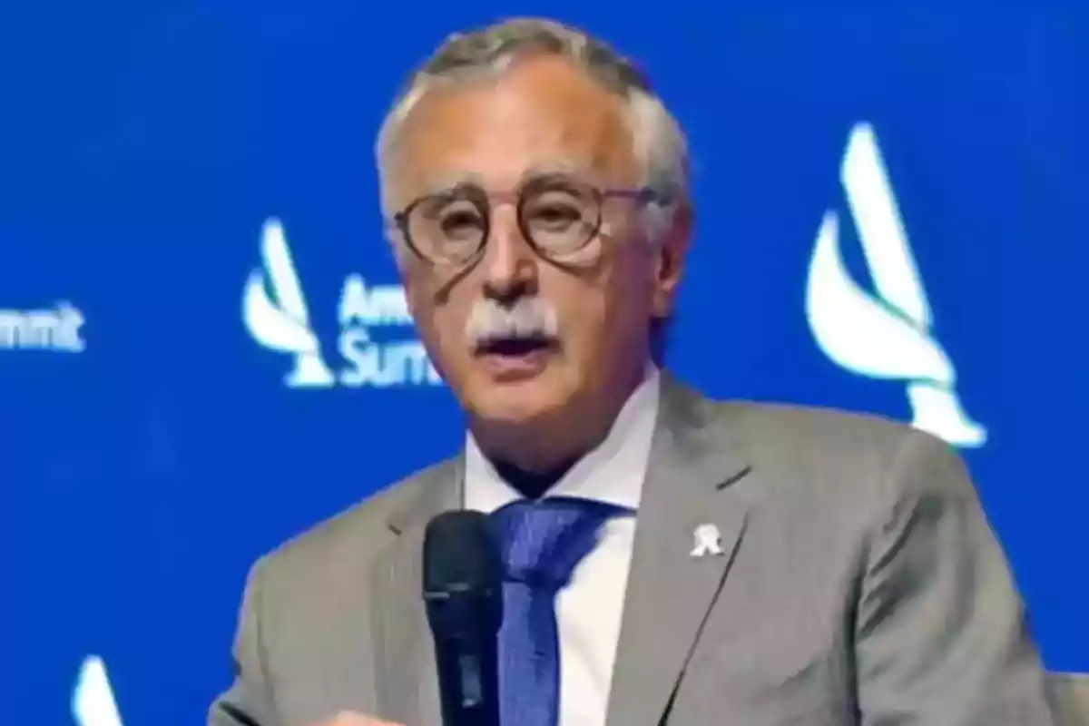 Hombre de cabello canoso y bigote con gafas y traje gris hablando en un evento con fondo azul Hombre de cabello canoso y bigote con gafas y traje gris hablando en un evento con fondo azul