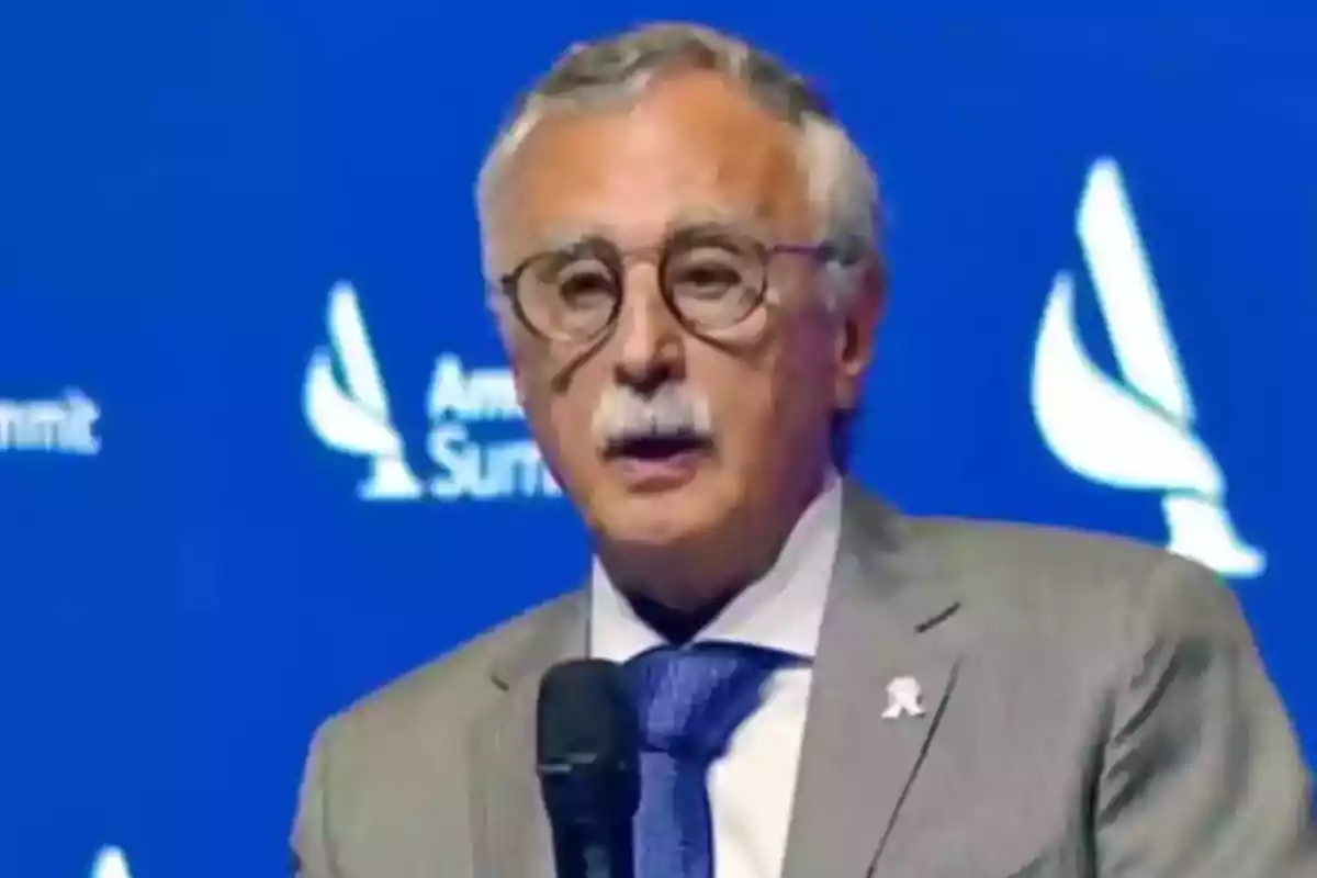 Hombre mayor con gafas y bigote hablando en un evento con fondo azul