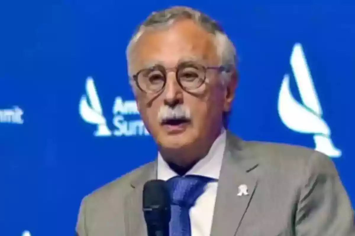 Hombre de cabello canoso y bigote usando gafas y traje gris hablando frente a un micrófono con fondo azul Hombre de cabello canoso y bigote usando gafas y traje gris hablando frente a un micrófono con fondo azul