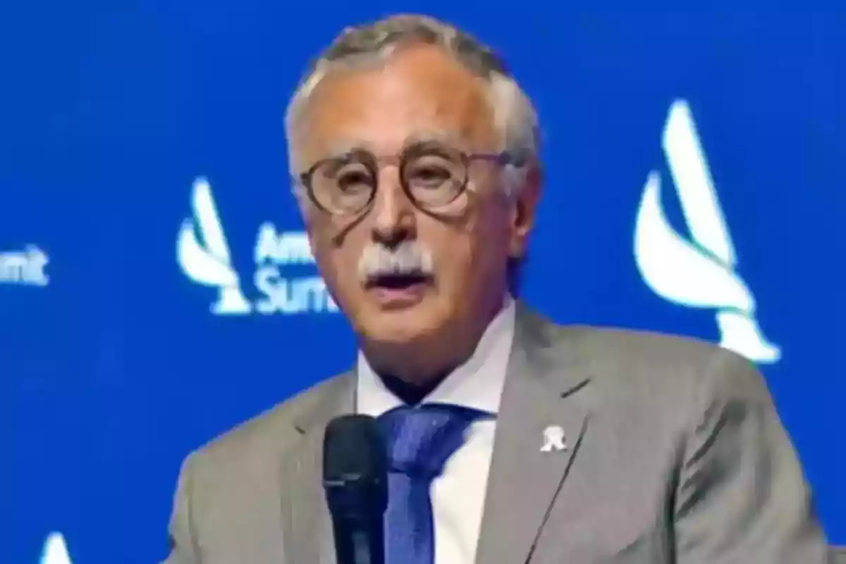 Hombre de cabello canoso y bigote con gafas hablando en un micrófono frente a un fondo azul con logotipos blancos Hombre de cabello canoso y bigote con gafas hablando en un micrófono frente a un fondo azul con logotipos blancos