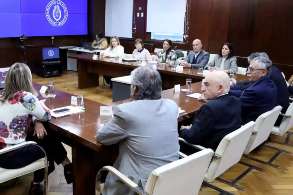 Varias personas sentadas alrededor de una mesa de reuniones en una sala con paredes de madera y una pantalla grande al fondo