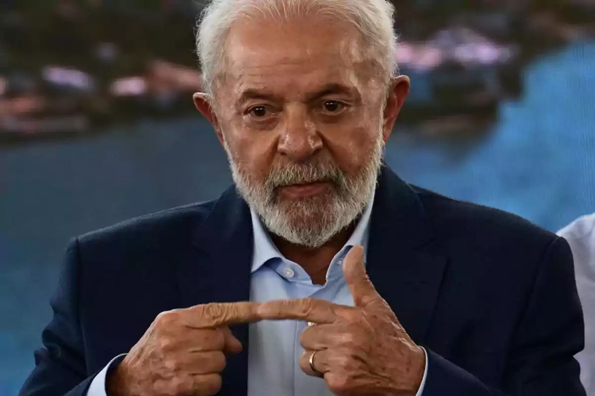 Hombre de cabello y barba canosos con saco azul marino y camisa celeste haciendo un gesto con las manos apuntando a su dedo índice Hombre de cabello y barba canosos con saco azul marino y camisa celeste haciendo un gesto con las manos apuntando a su dedo índice