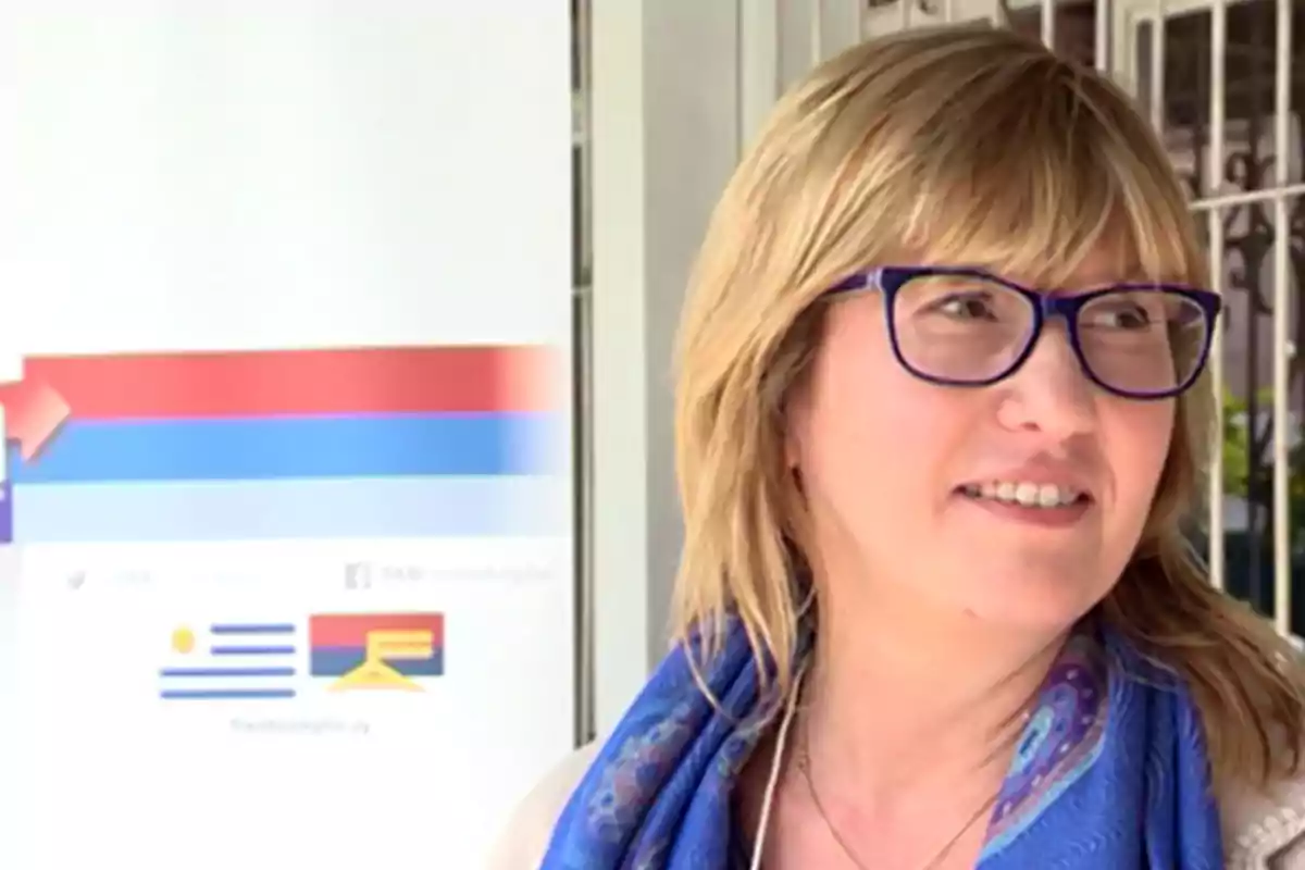 Mujer rubia con gafas y bufanda azul sonriendo frente a un cartel con los colores de la bandera de Uruguay Mujer rubia con gafas y bufanda azul sonriendo frente a un cartel con los colores de la bandera de Uruguay
