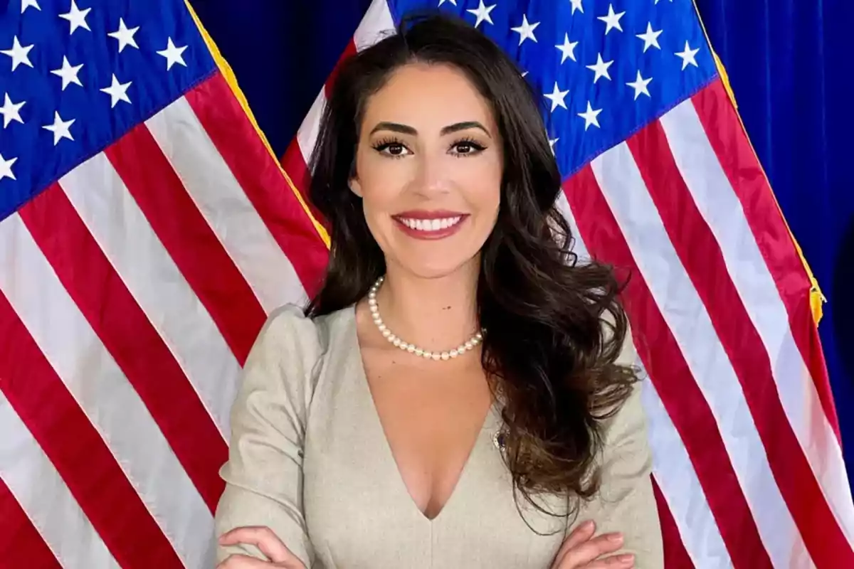 Smiling woman with long dark hair and a pearl necklace, formally dressed with her arms crossed, posing in front of two United States flags Smiling woman with long dark hair and a pearl necklace, formally dressed with her arms crossed, posing in front of two United States flags