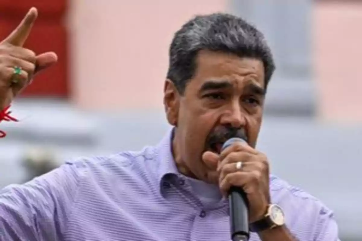 Hombre de cabello canoso y bigote hablando en un micrófono y levantando una mano mientras usa una camisa de cuadros morada Hombre de cabello canoso y bigote hablando en un micrófono y levantando una mano mientras usa una camisa de cuadros morada