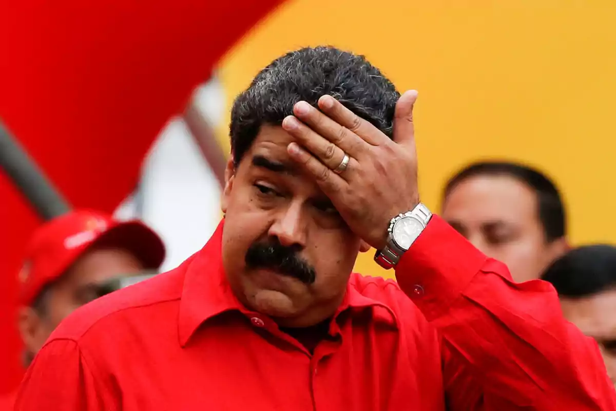 Man in a red jersey puts his hand to his forehead in a gesture of concern while surrounded by people in a colorful setting.