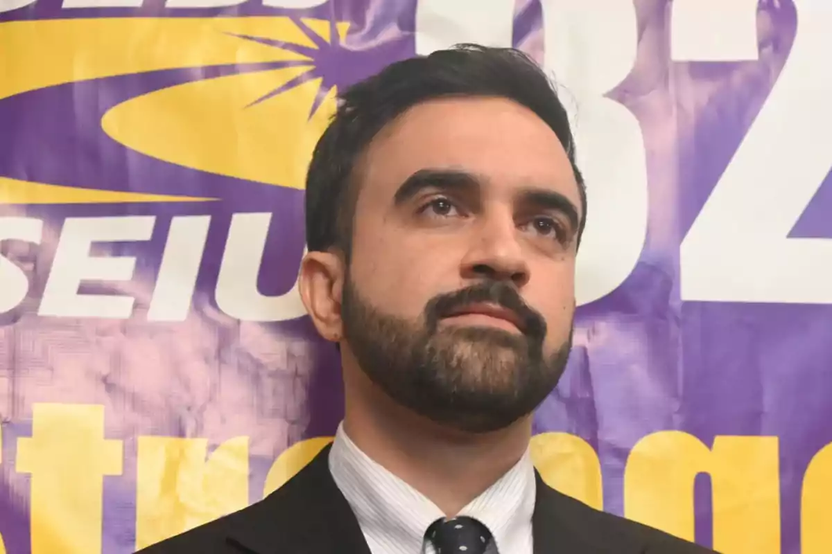 Dark-haired, bearded man in a suit and tie in front of a purple and yellow sign with large letters