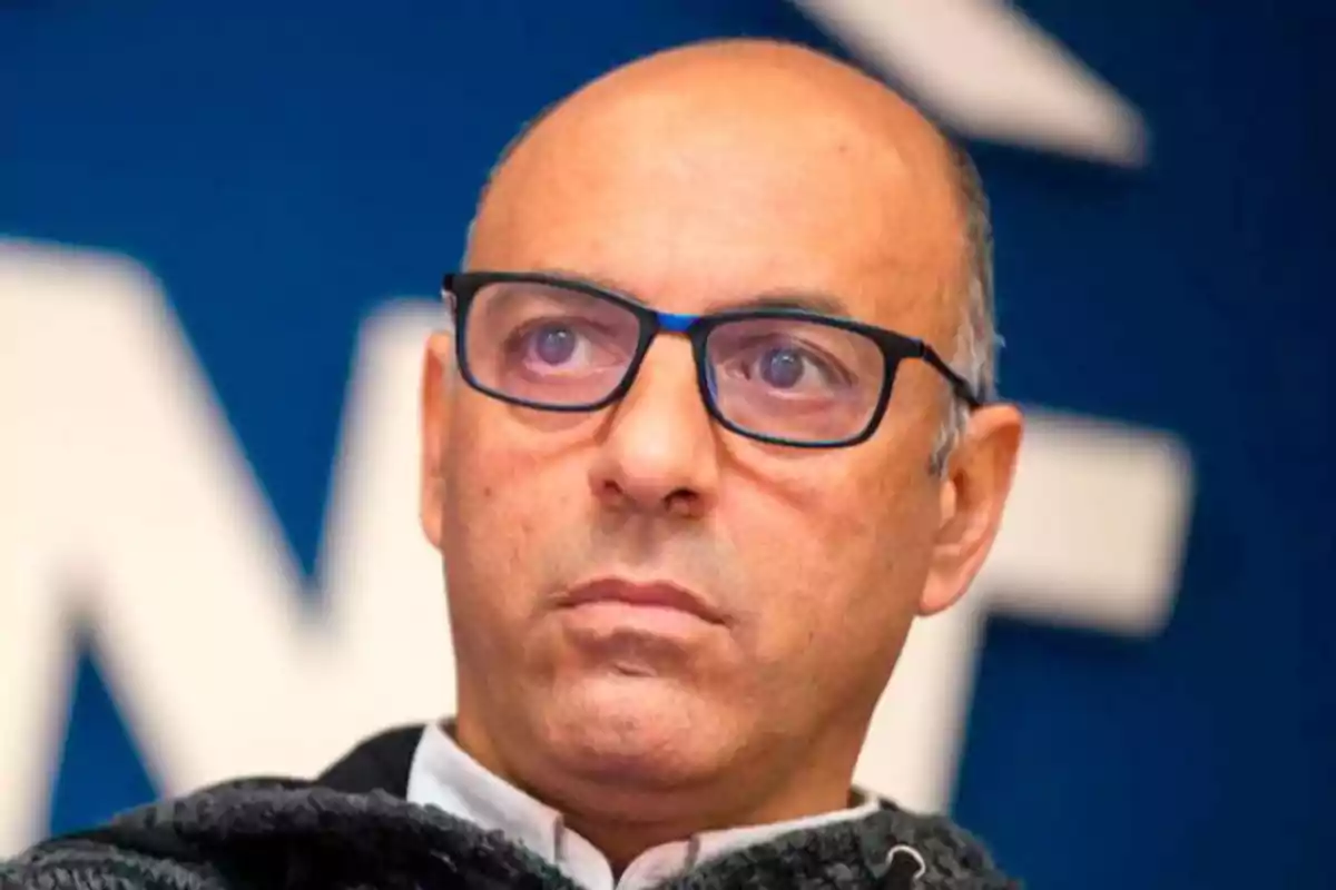 Hombre calvo con gafas negras y expresión seria frente a un fondo azul desenfocado