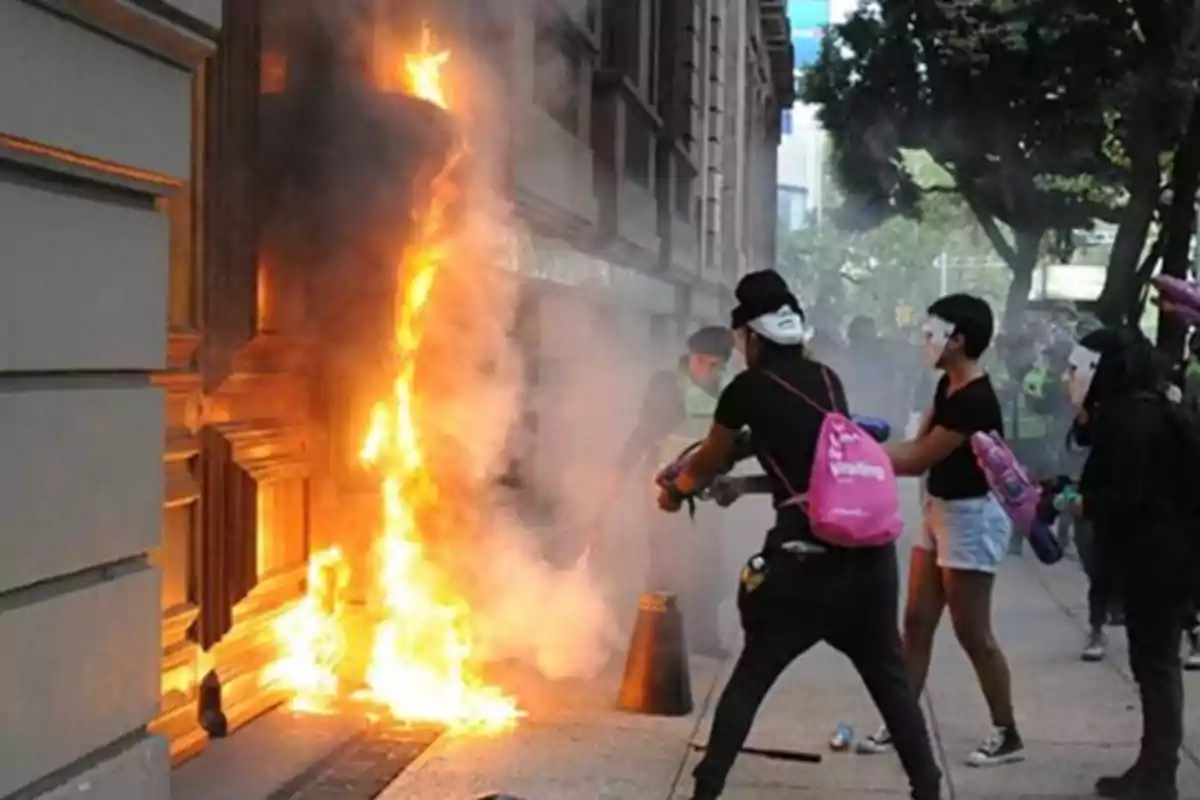 People wearing masks and backpacks watch a fire at the entrance of a building as flames and smoke rise up the facade. People wearing masks and backpacks watch a fire at the entrance of a building as flames and smoke rise up the facade.