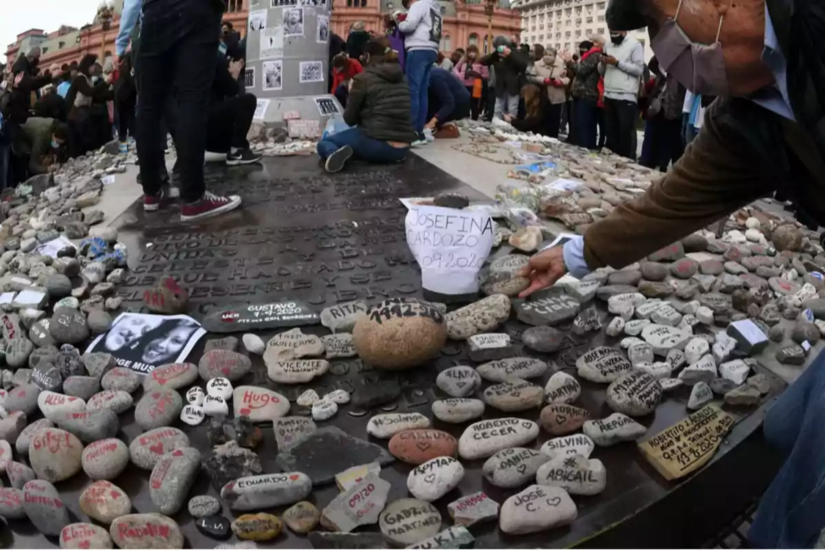 Personas reunidas en una plaza dejando piedras con nombres escritos en memoria de seres queridos frente a un monumento rodeado de mensajes y fotografías Personas reunidas en una plaza dejando piedras con nombres escritos en memoria de seres queridos frente a un monumento rodeado de mensajes y fotografías