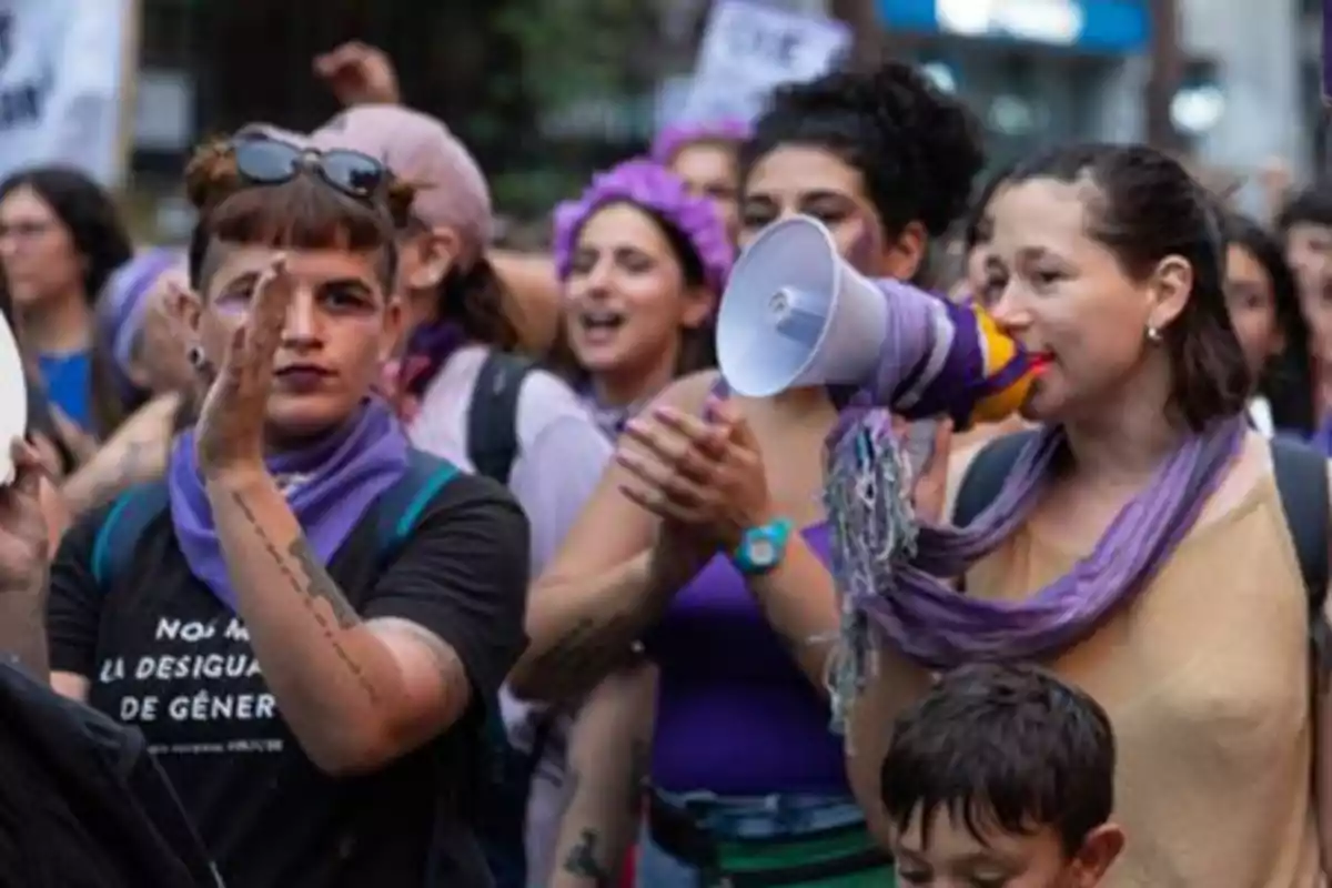 Women marching in a feminist demonstration with purple scarves and clothing, one of them using a megaphone to speak