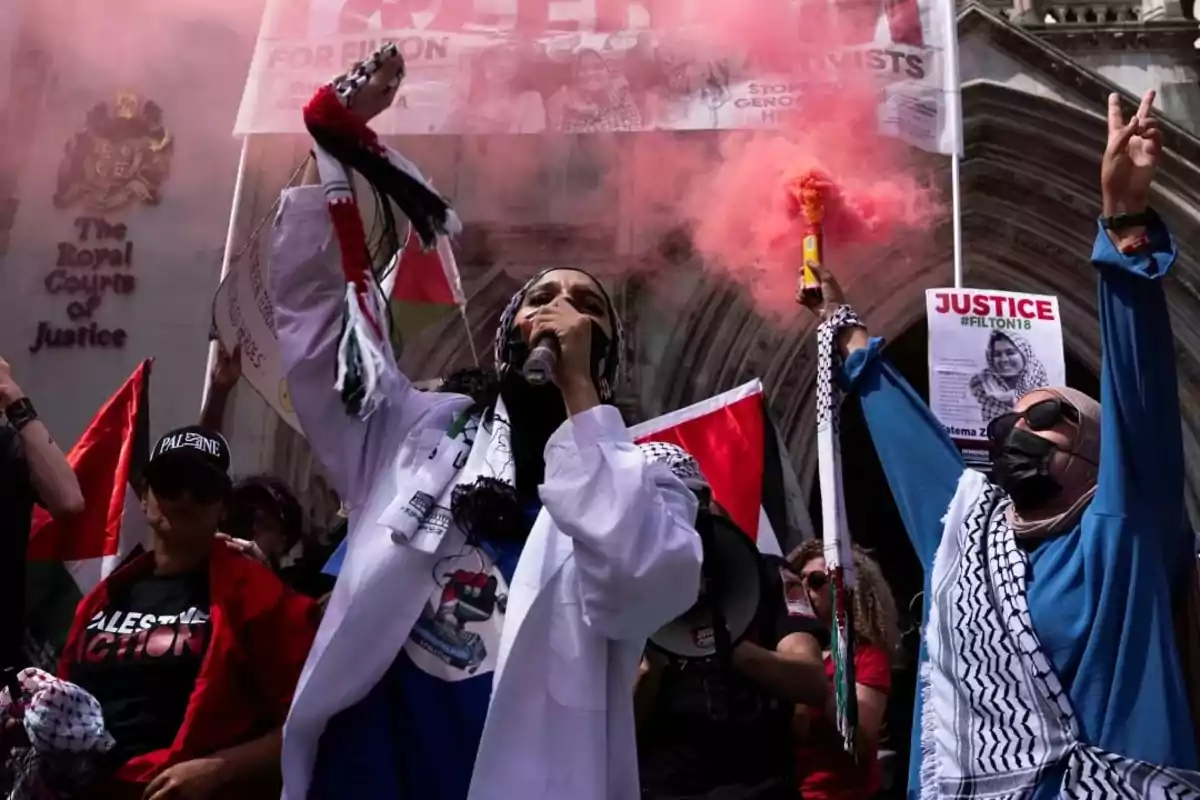 People showing in front of a building with signs, flags, and red smoke, showing gestures of protest and solidarity