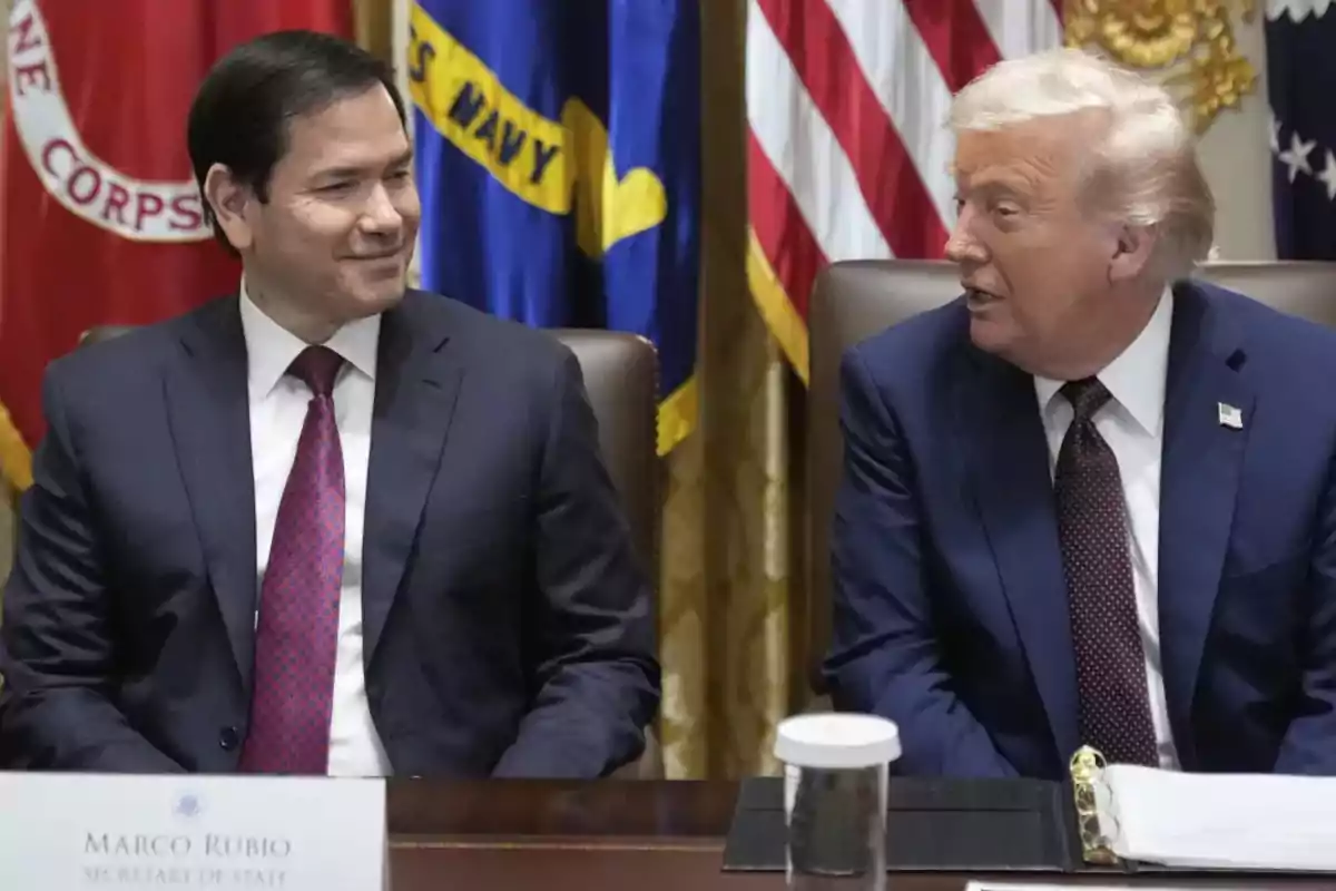Two men in suits are sitting at a conference table talking, with flags in the background.
