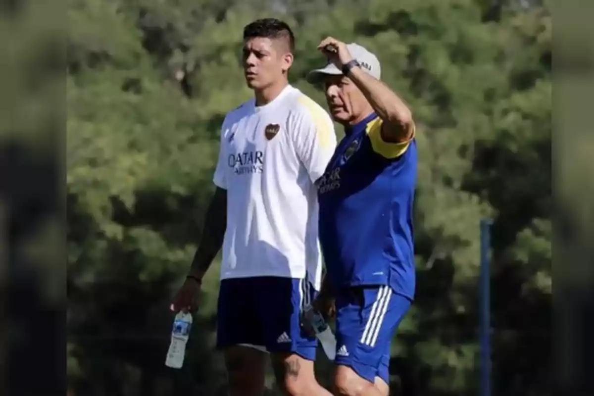 Dos personas con ropa deportiva de Boca Juniors están de pie en un campo de entrenamiento rodeado de árboles, ambas sostienen botellas de agua y parecen estar conversando o analizando algo. Dos personas con ropa deportiva de Boca Juniors están de pie en un campo de entrenamiento rodeado de árboles, ambas sostienen botellas de agua y parecen estar conversando o analizando algo.