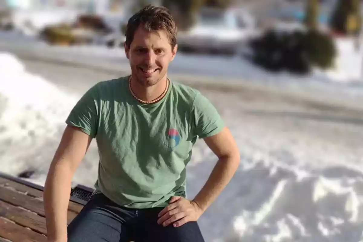 Hombre joven con camiseta verde sentado al aire libre en un entorno nevado sonriendo a la cámara Hombre joven con camiseta verde sentado al aire libre en un entorno nevado sonriendo a la cámara