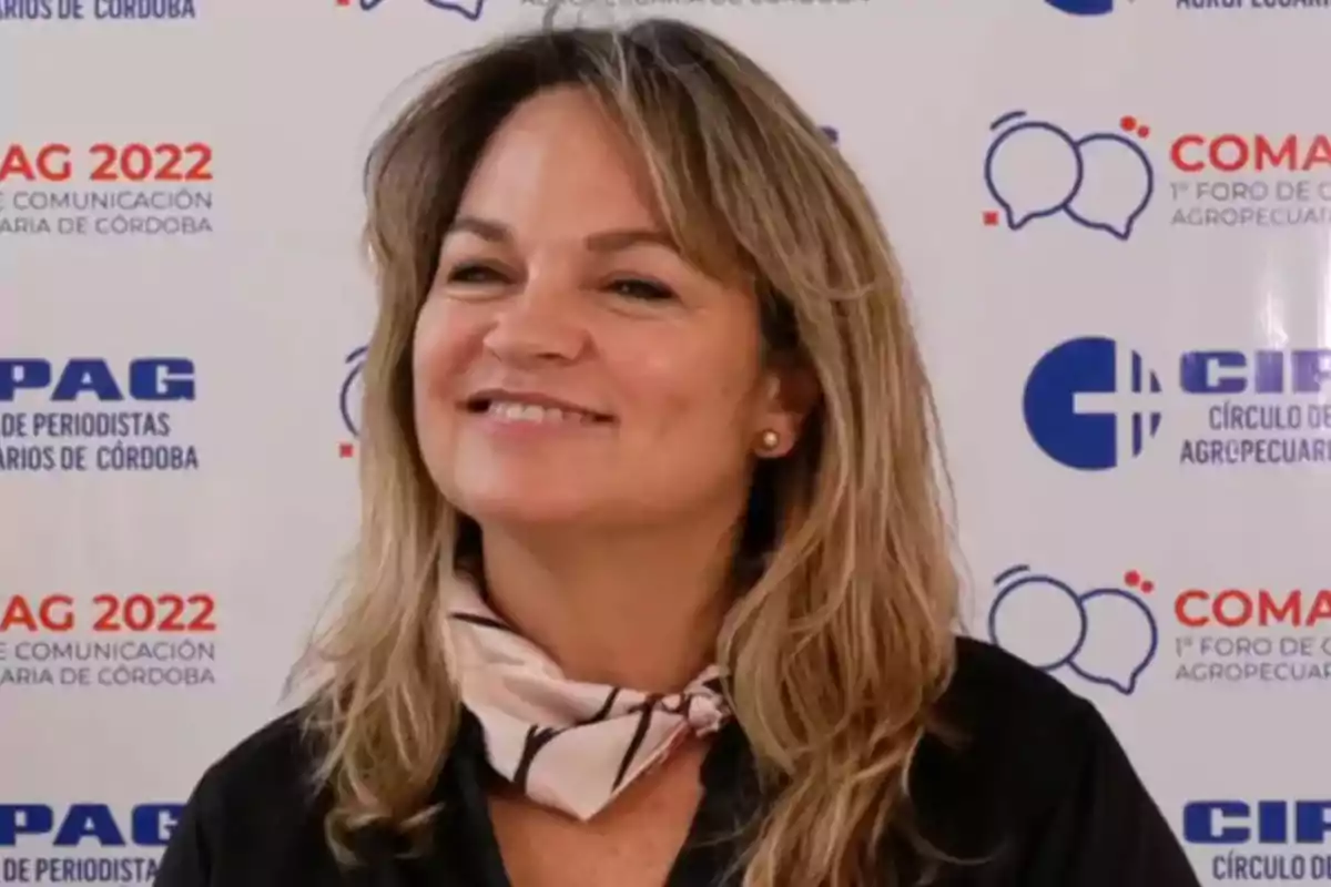 Smiling woman with blonde hair and a light scarf around her neck posing in front of a backdrop with logos from an agricultural communication event
