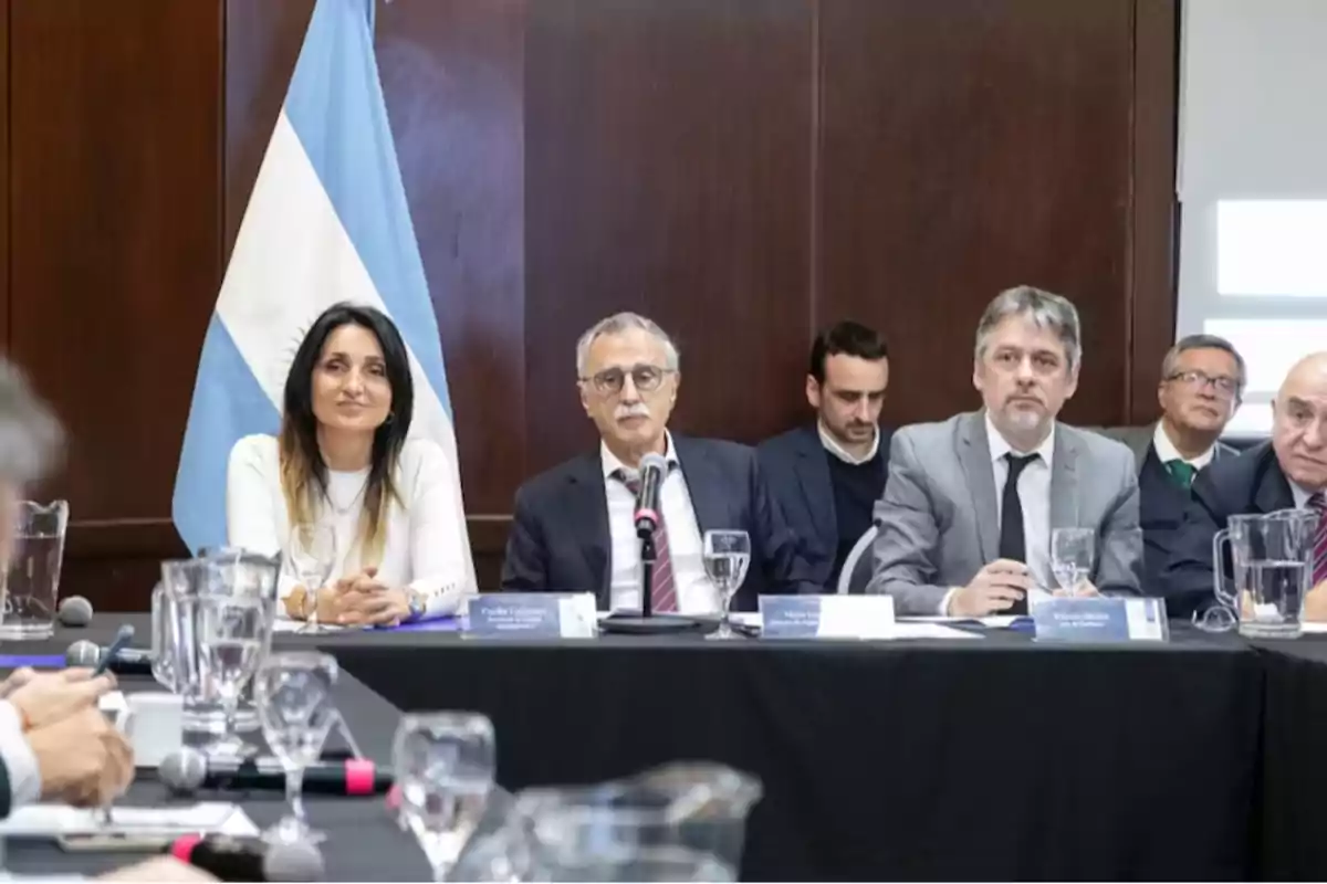 Varias personas sentadas en una mesa de reunión formal con una bandera de Argentina de fondo
