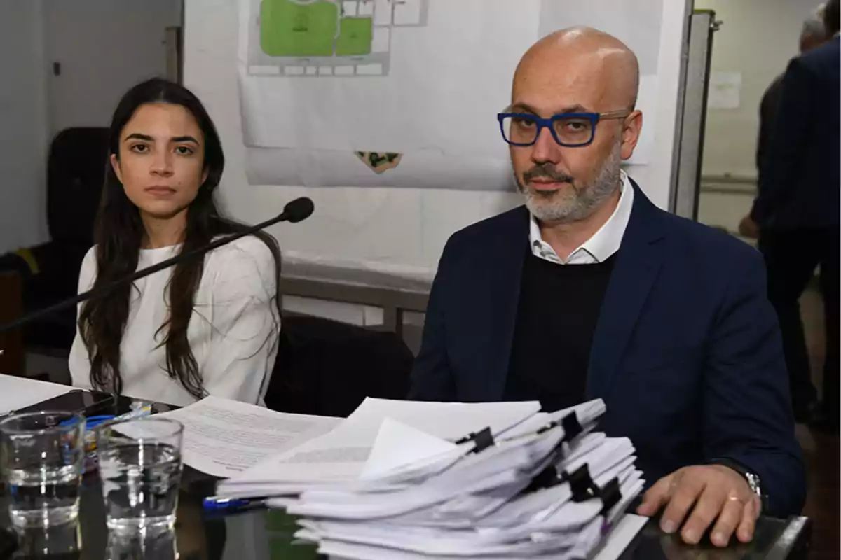 Dos personas sentadas en una mesa con documentos y vasos de agua, una mujer de cabello largo y un hombre con gafas y barba, en un ambiente de oficina o reunión.