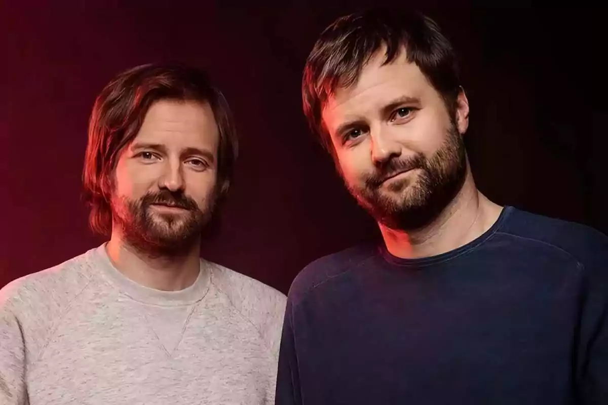 Two men with beards and brown hair pose together in front of a dark background with reddish lighting. Two men with beards and brown hair pose together in front of a dark background with reddish lighting.