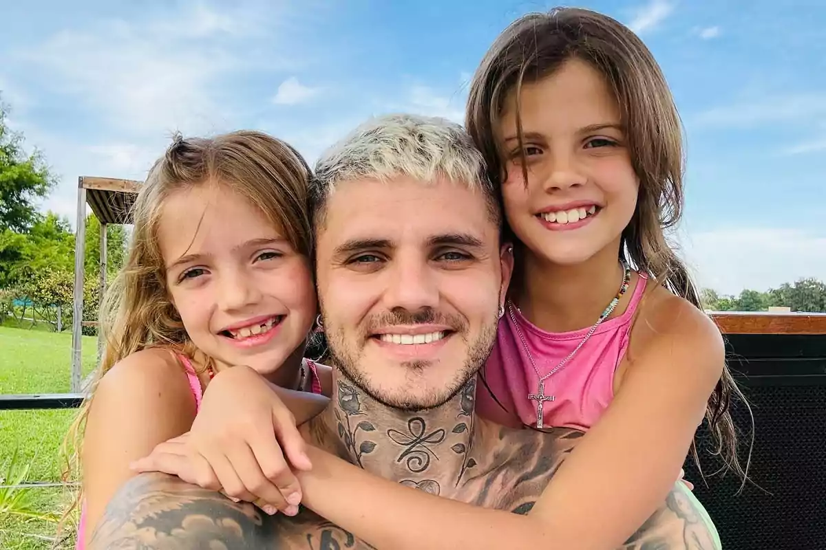 Un hombre sonriente con cabello corto y rubio posa abrazado por dos niñas pequeñas que también sonríen, todos al aire libre en un día soleado Un hombre sonriente con cabello corto y rubio posa abrazado por dos niñas pequeñas que también sonríen, todos al aire libre en un día soleado