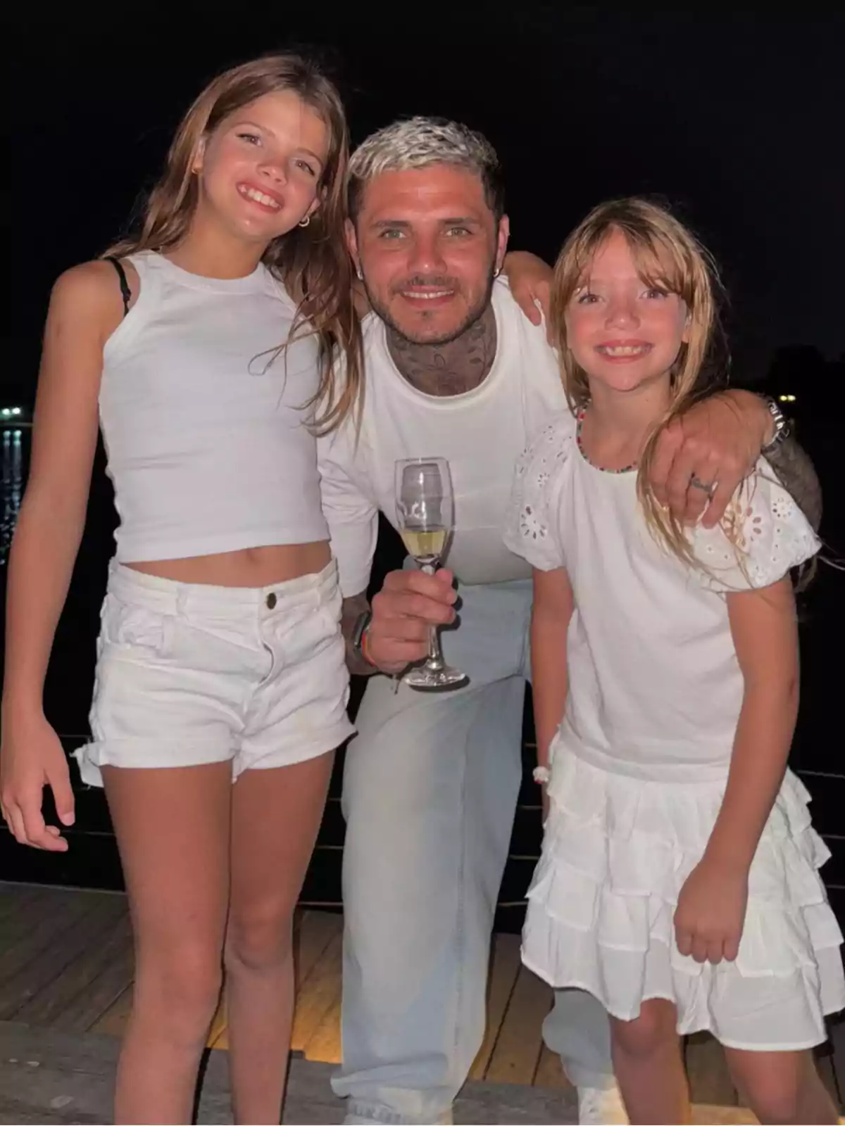 Tres personas sonríen y posan juntas en una terraza de noche, todas vestidas de blanco, mientras una de ellas sostiene una copa Tres personas sonríen y posan juntas en una terraza de noche, todas vestidas de blanco, mientras una de ellas sostiene una copa