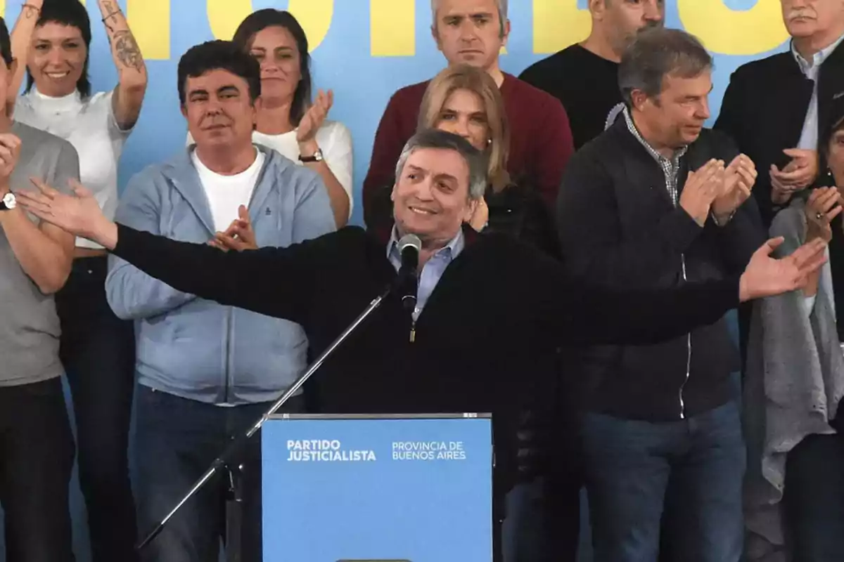 Un hombre sonriente con los brazos abiertos habla en un podio frente a un grupo de personas que lo aplauden Un hombre sonriente con los brazos abiertos habla en un podio frente a un grupo de personas que lo aplauden