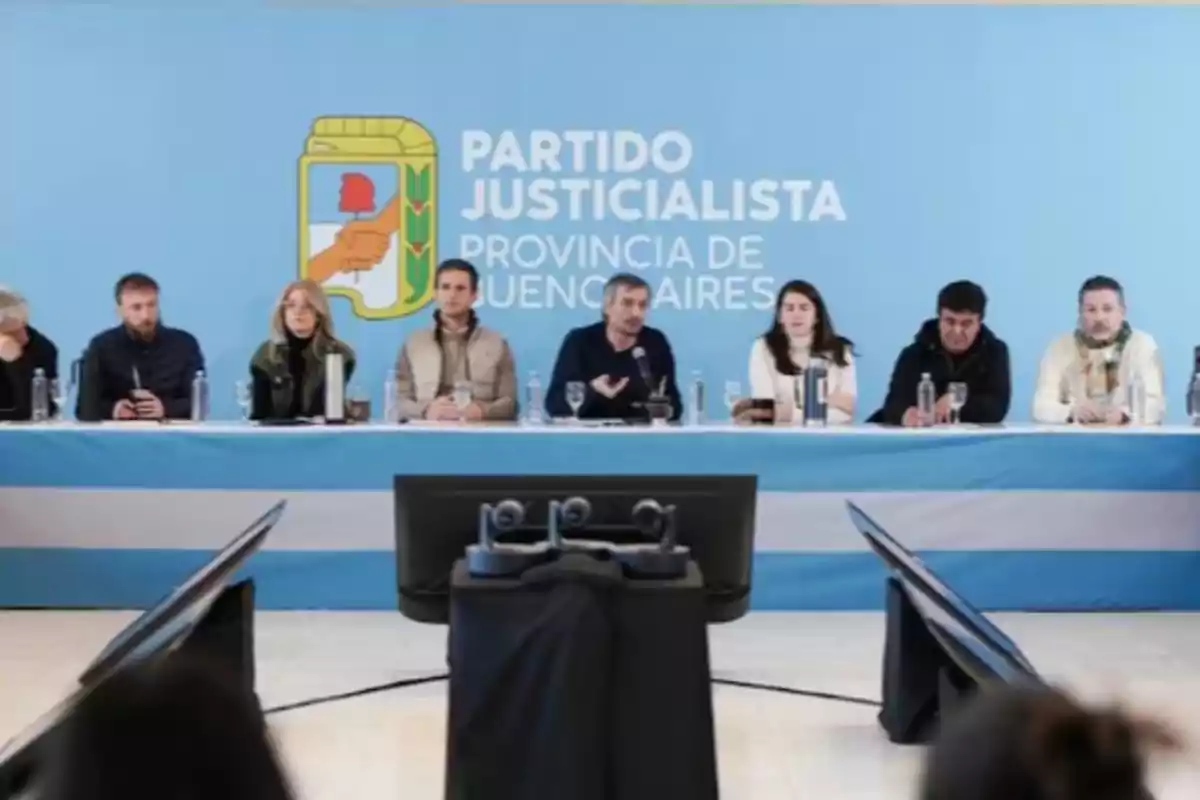 Varias personas sentadas en una mesa larga frente a un fondo celeste con el logo y nombre del Partido Justicialista de la provincia de Buenos Aires