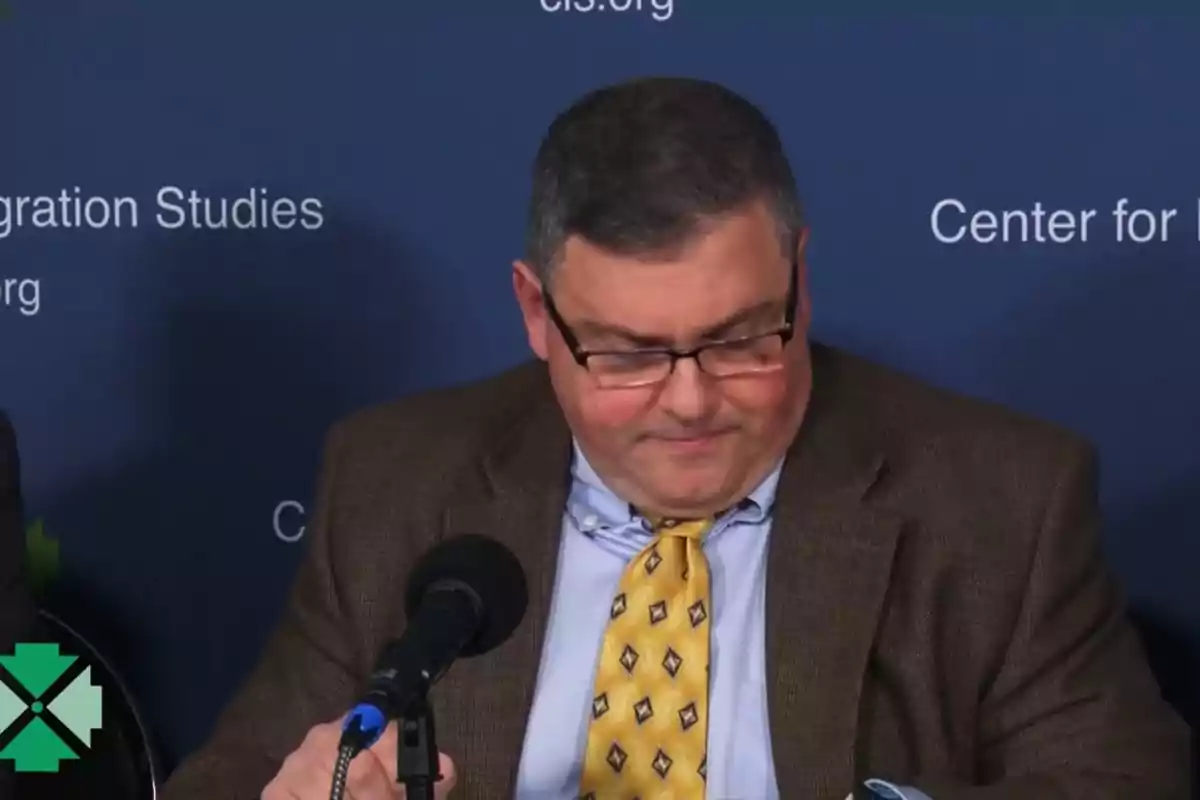 Hombre con gafas y corbata amarilla sentado frente a un micrófono en una conferencia con fondo azul Hombre con gafas y corbata amarilla sentado frente a un micrófono en una conferencia con fondo azul
